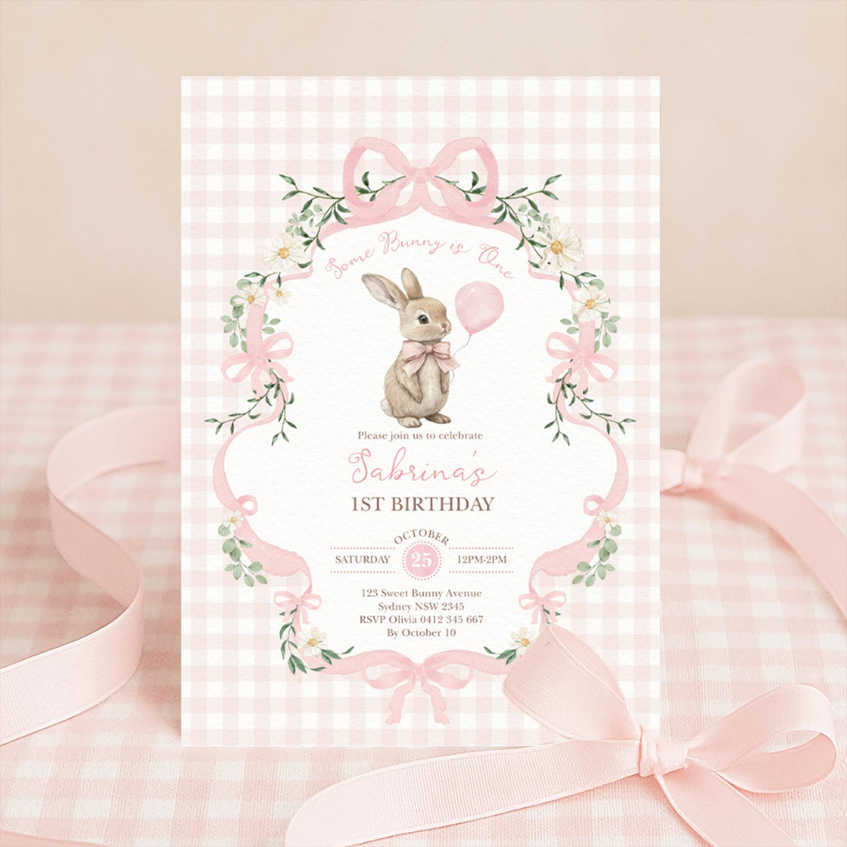Invitation standing upright with a pale pink ribbon behind it on pink gingham cloth