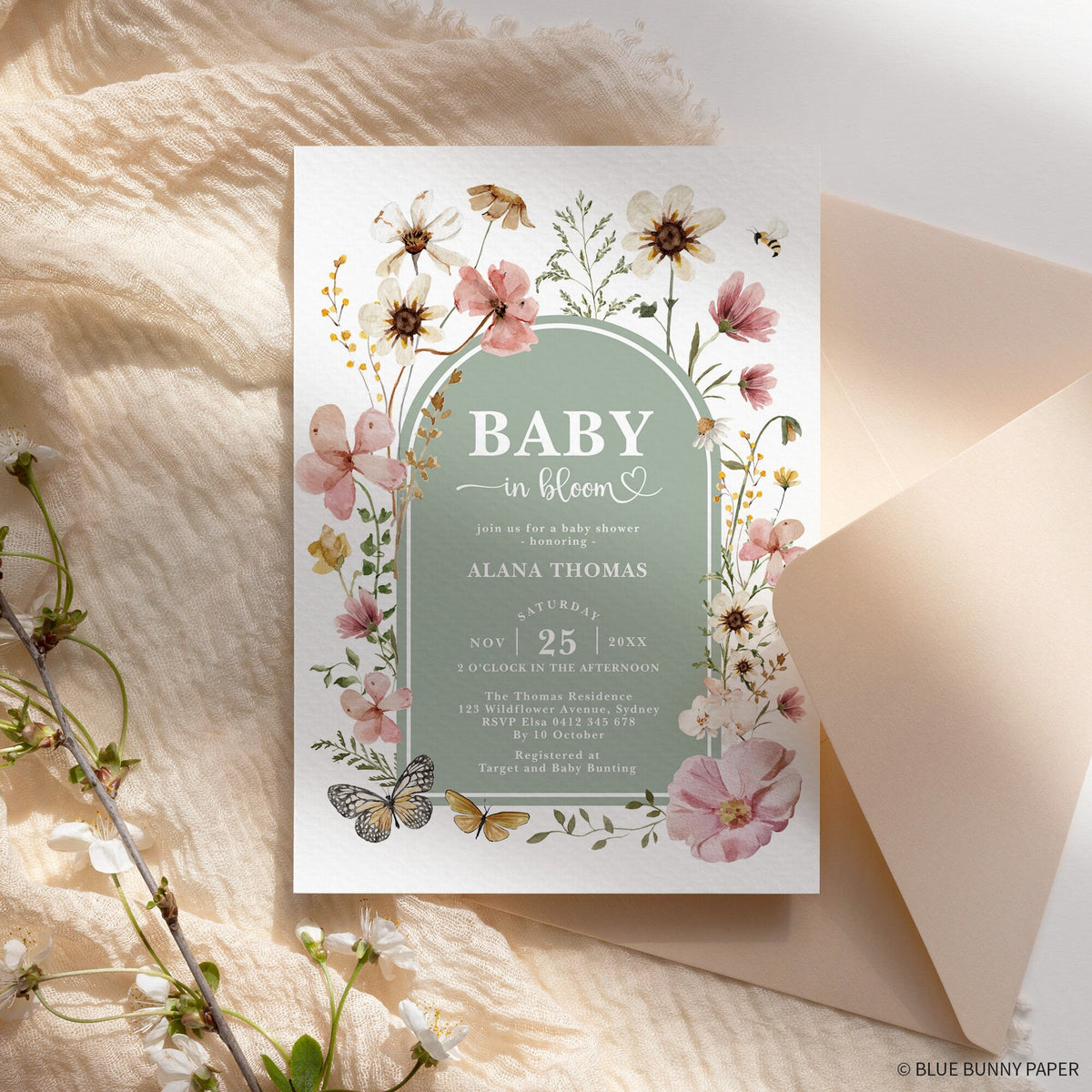 Sage wildflower baby shower invitation against blush envelopes and dried grass