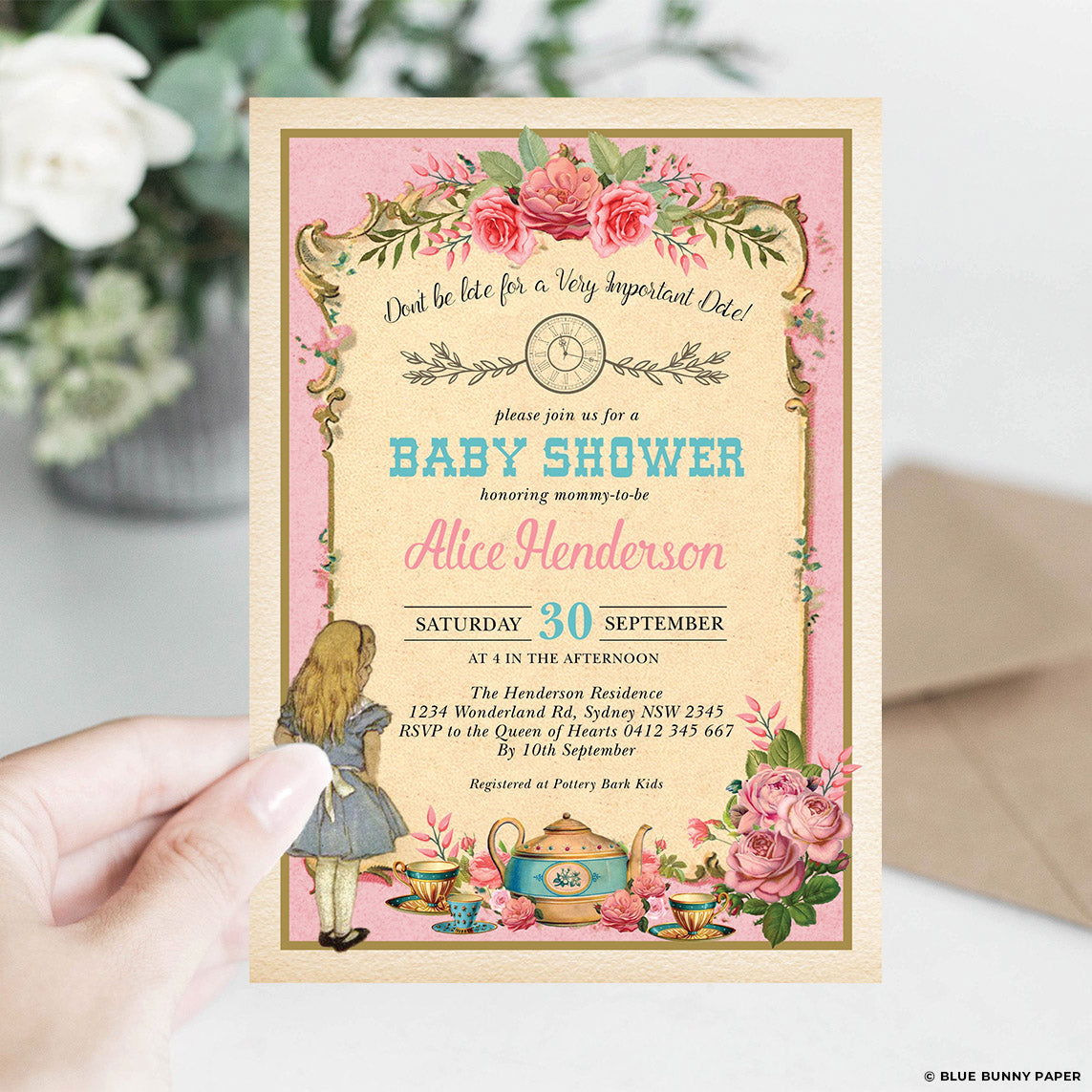 Pink baby shower invitation held in a hand in front of a soft white flower, gold border in view