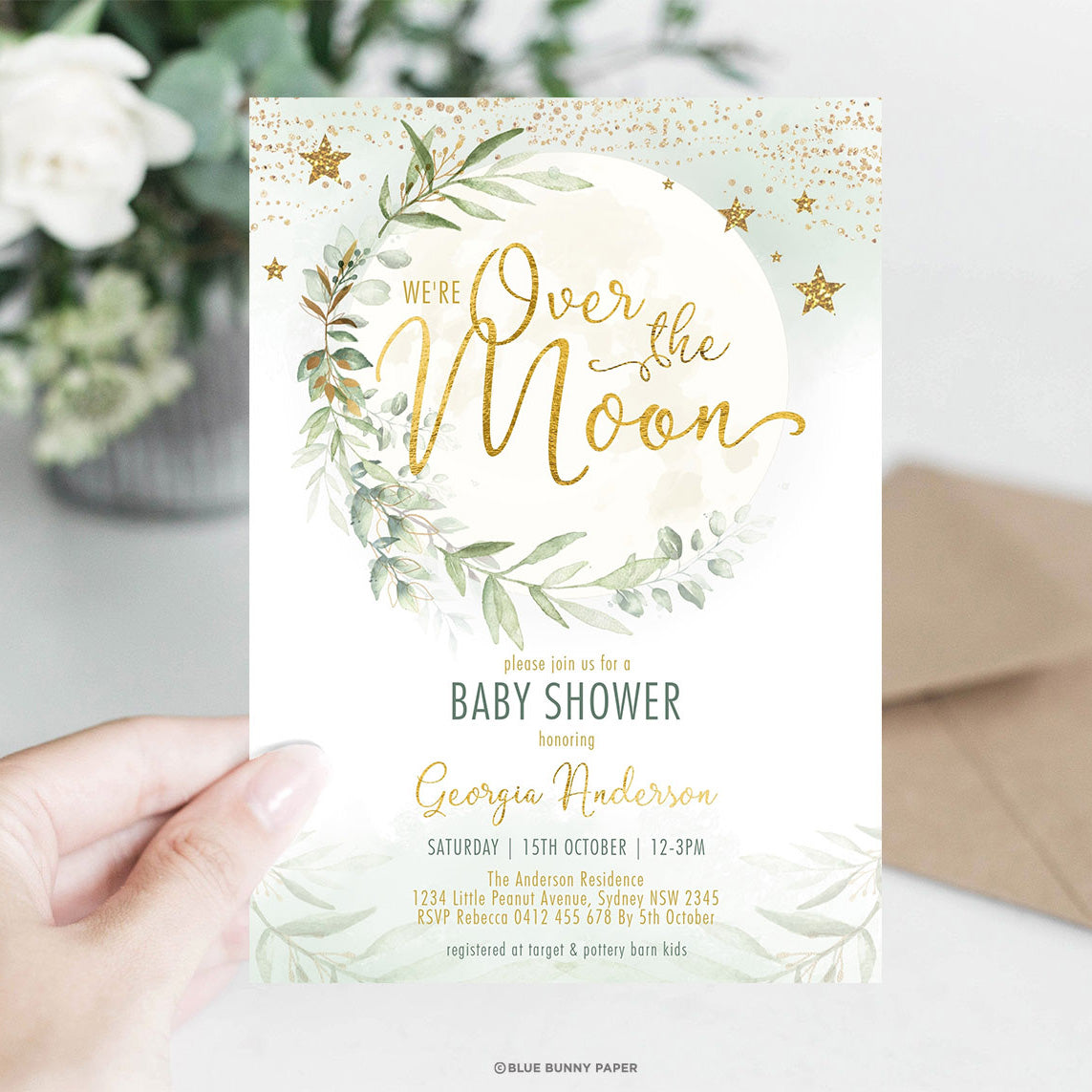 Gold foil Over the Moon baby shower invitation held in a hand against a blurred kraft envelope