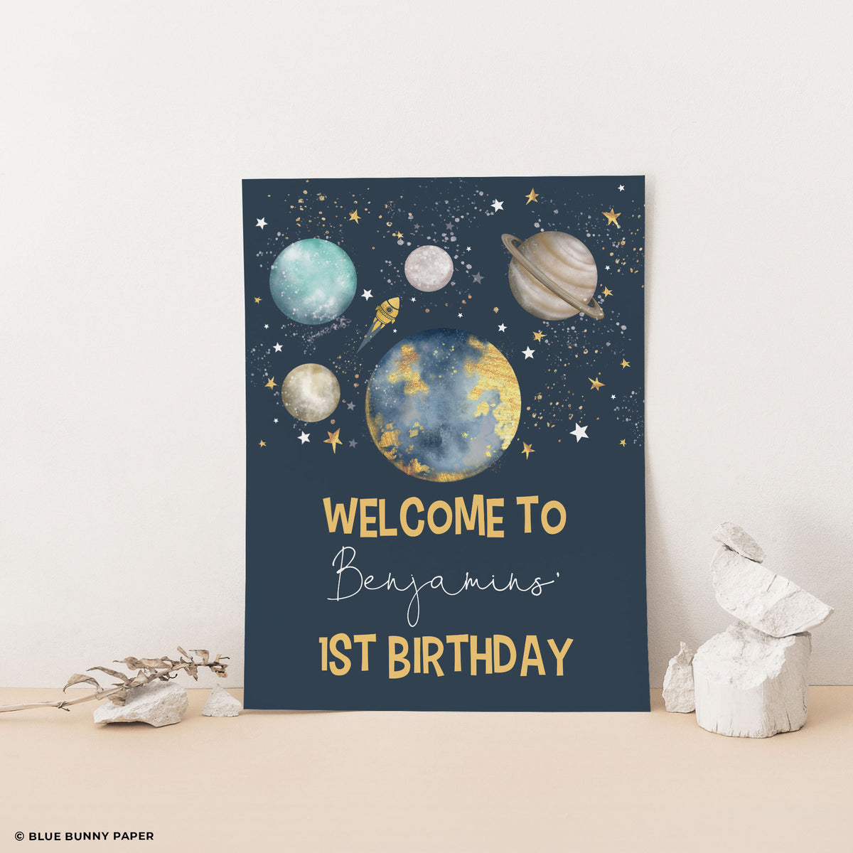 Navy galaxy welcome sign reading Welcome to Benjamin's 1st Birthday, propped up with white stones